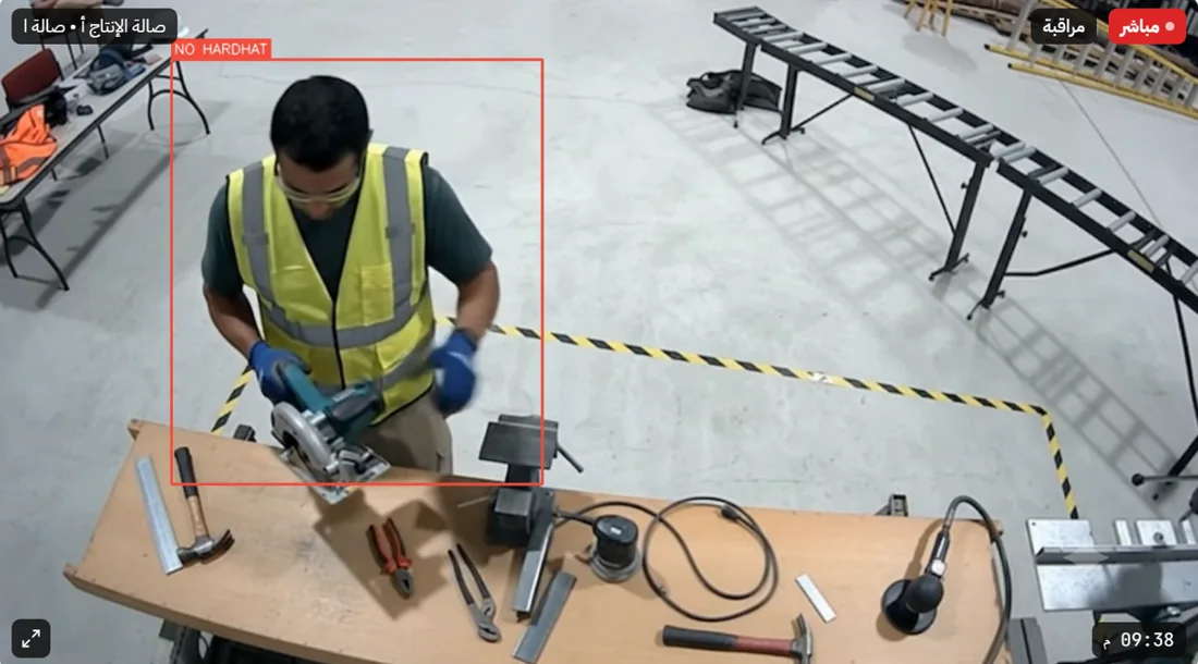 كاميرا داخل صالة إنتاج: إطار أحمر حول عاملٍ يشغّل منشاراً بلا خوذة، مع تسمية «NO HARDHAT».